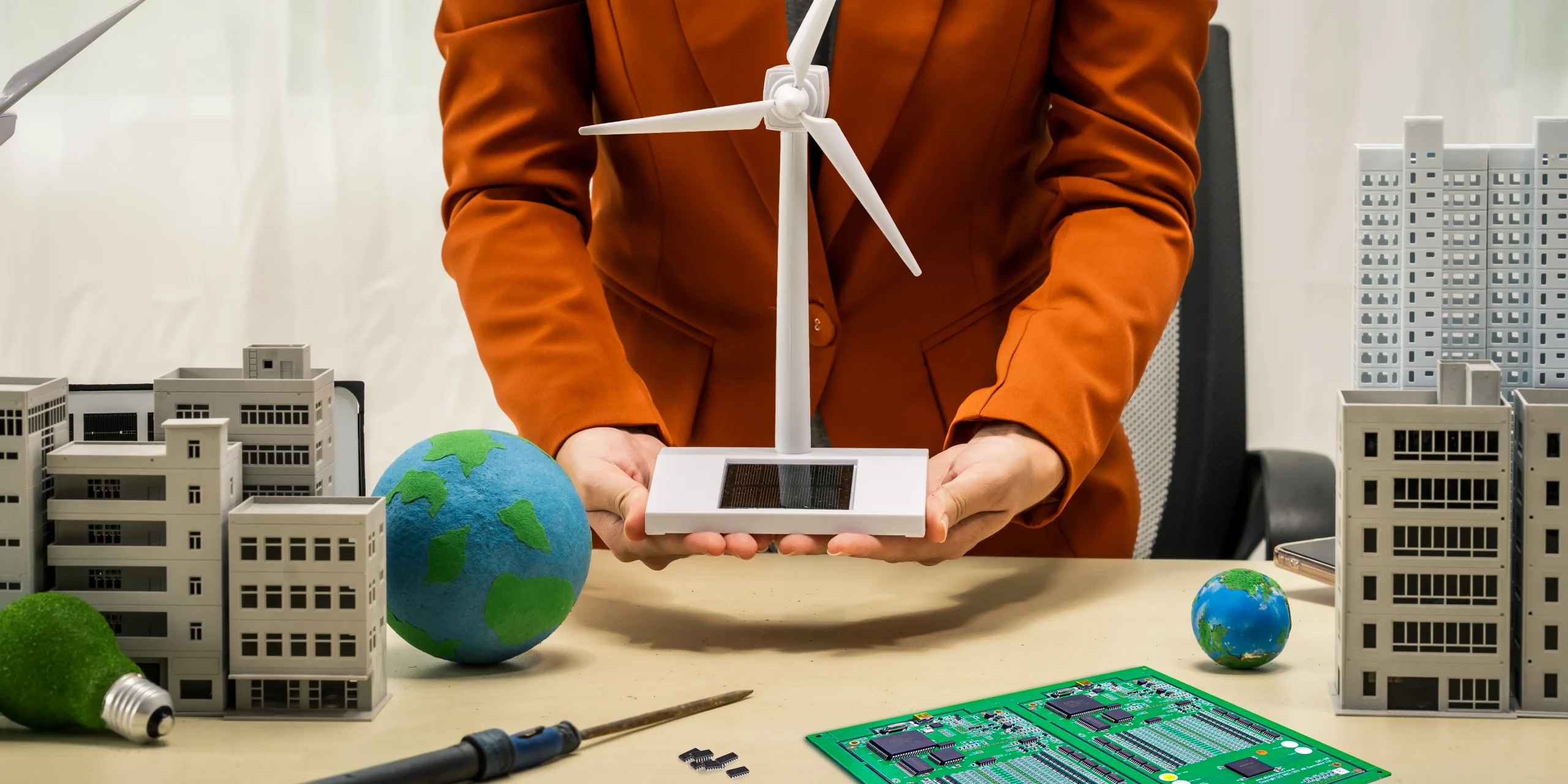 Business woman holding a model wind turbine with a repaired circuit board/PCB on the desk next to soldering iron and some integrated circuit chips. A metaphor for how electronics repair benefits renewable energy sustainability.
